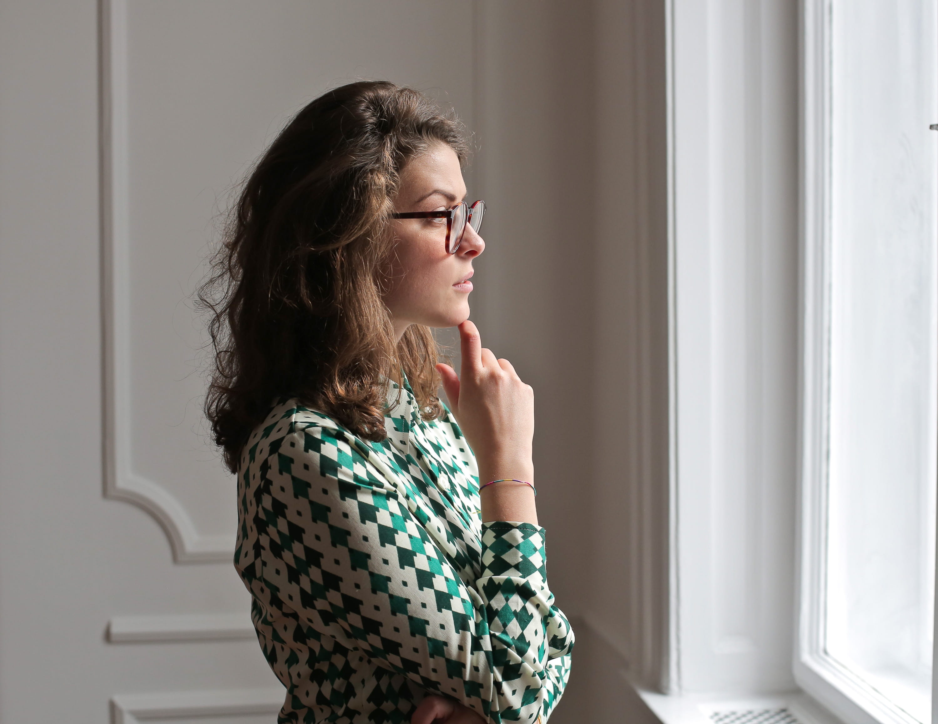 Young woman wearing printed collared shirt and spectacles keeping hand on chin and looking away while standing near window