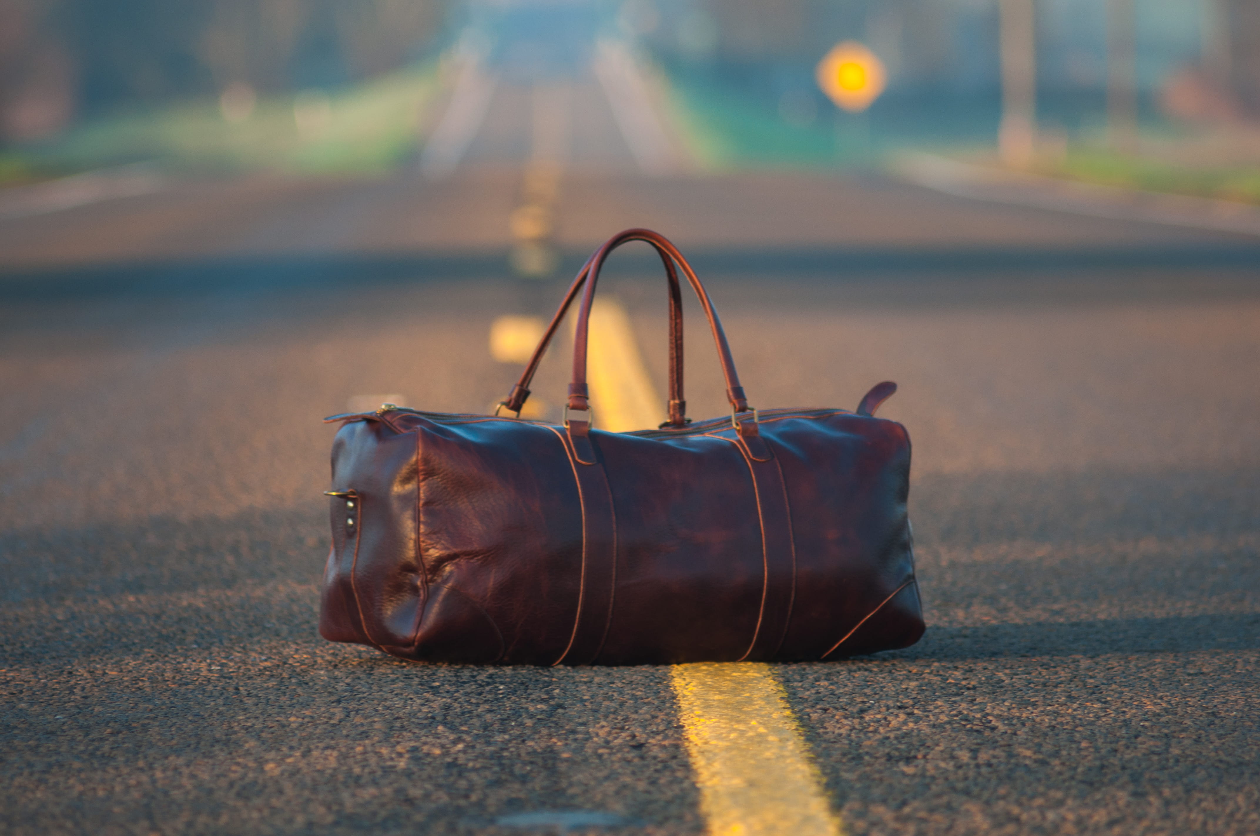 brown leather duffel bag in middle on gray asphalt road, focus on foreground