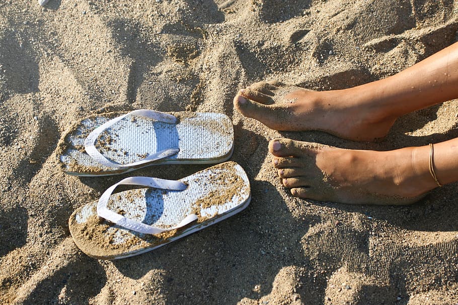 Relaxed female feet with an anklet on the left foot on the sand with slippers
