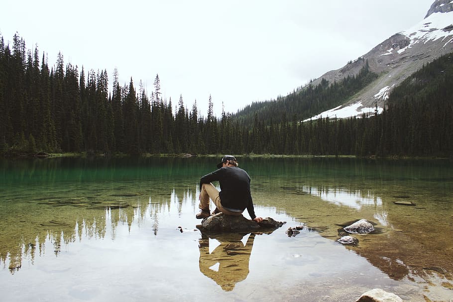 man wearing sweater sitting on stone by calm lake with trees on shore fronting snow capped mountain at daytime HD wallpaper