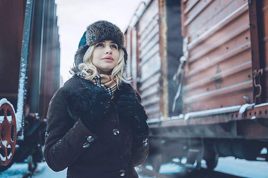 woman wearing brown and black fur-lined coat and knit cap near train cars during snowy daytime