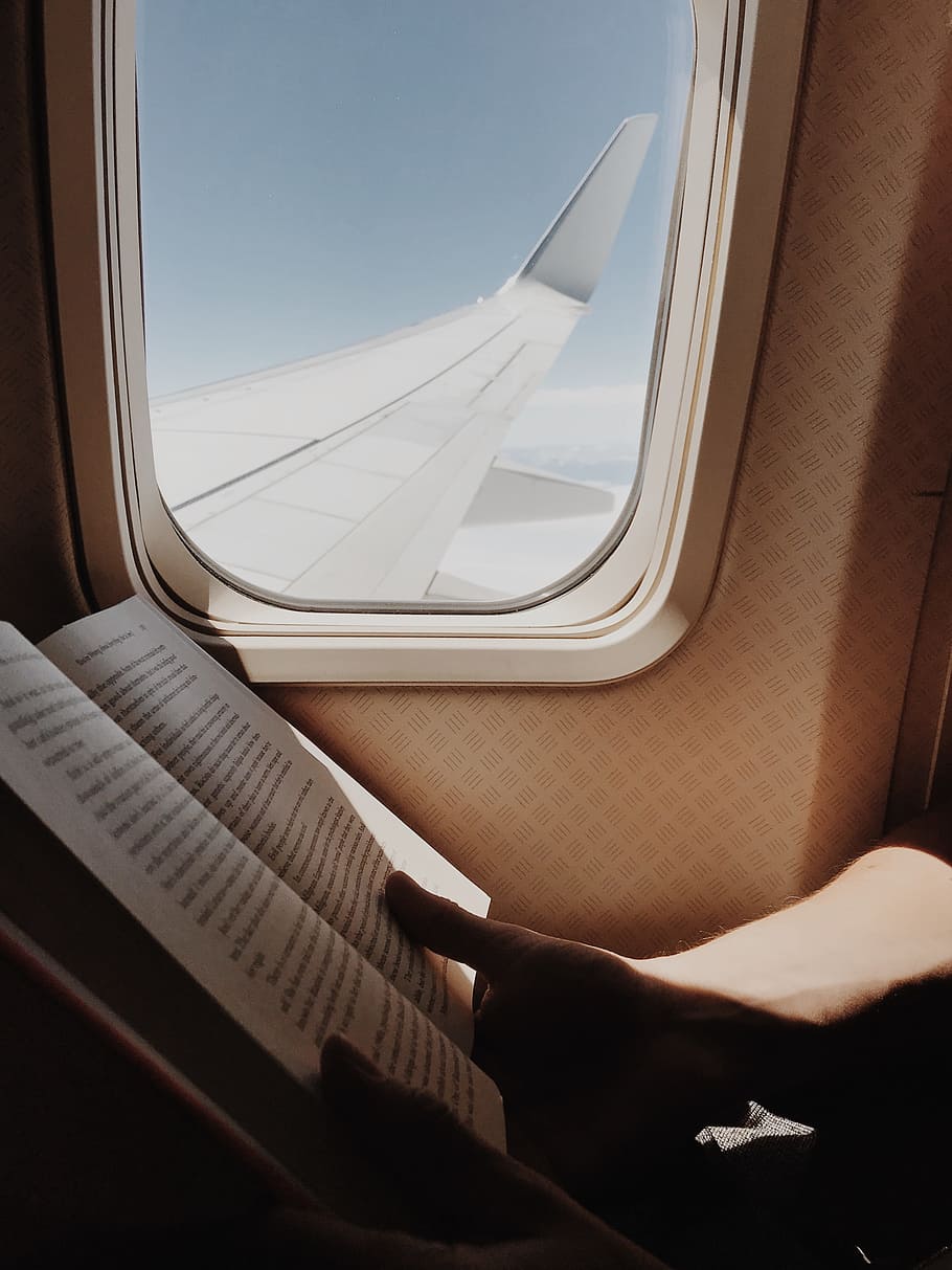 person reading book beside airplane window, wing, hold, hand