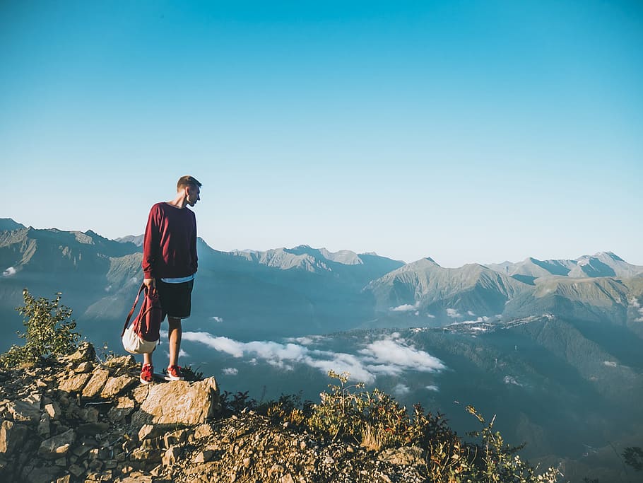Man in Red Sweatshirt and Black Shorts Standing on Large Brown Rock on Top of a Mountain