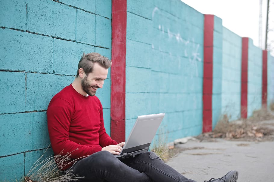 A young bearded man wearing a cherry red pullover sitting against a boundary wall smilingly looking at his laptop