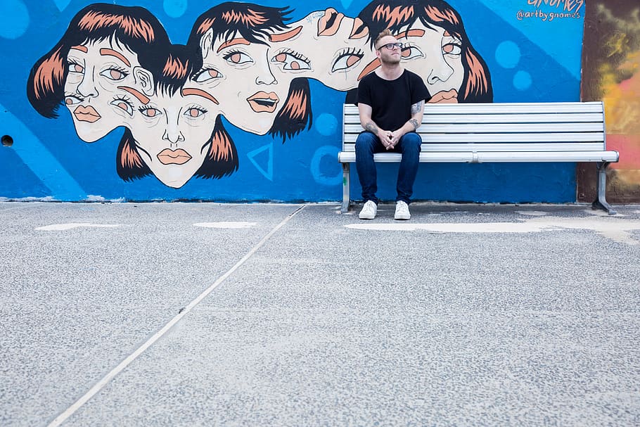 man sitting on white bench in front of wall with painting, clothing