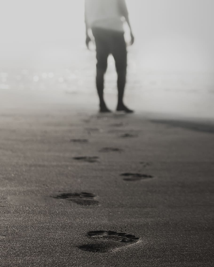 grayscale photography of man walking on sand leaving footprints