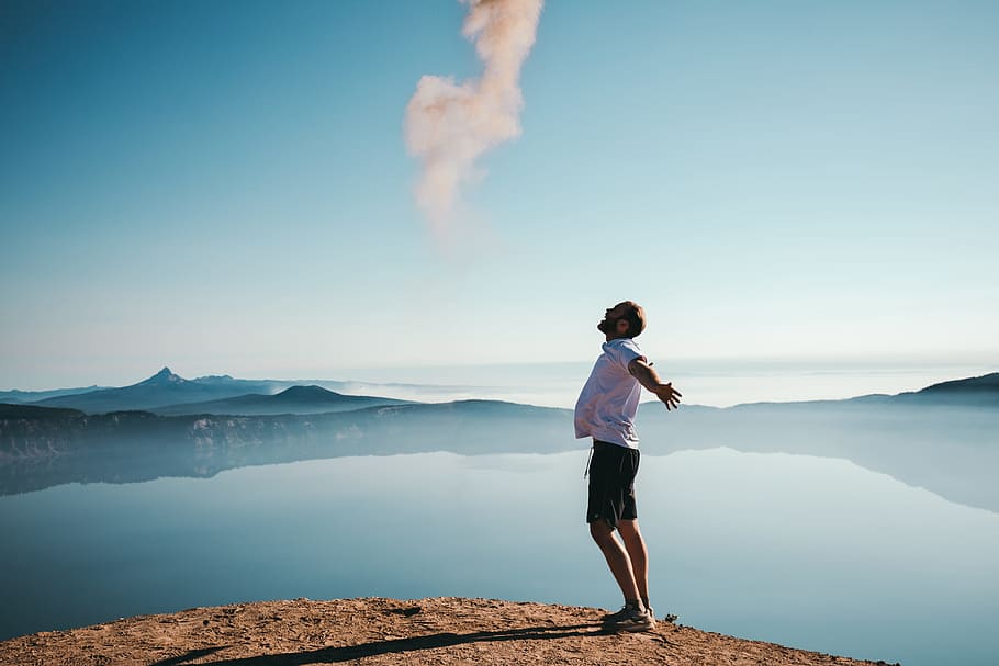 man standing on sand while spreading arms beside calm body of water HD wallpaper