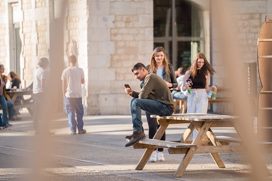 Man in Grey Long-sleeved Shirt and Blue Denim Jeans Sits on Picnic Table Near Women Walking