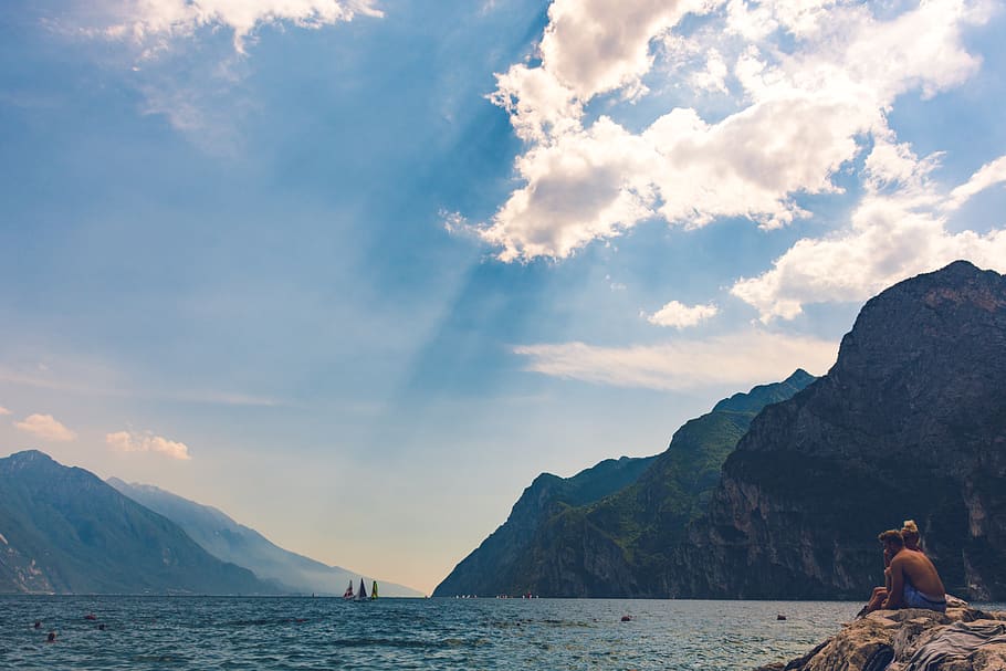 Man and Woman Sitting on Stone Facing Body of Water and Island HD wallpaper
