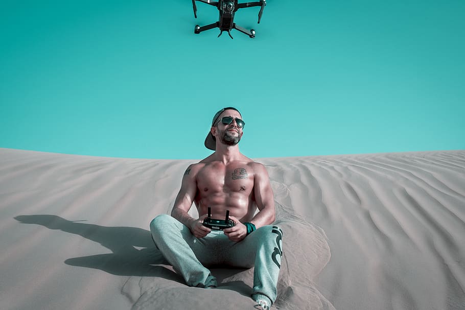 Man Sitting on Sand Controlling Drone, cap, controller, daylight HD wallpaper