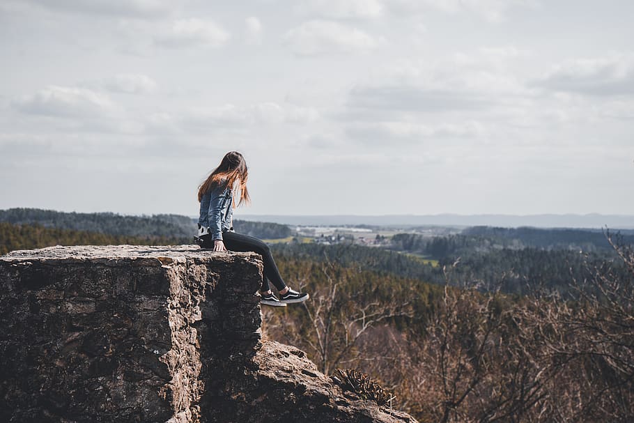 woman sitting on cliff's edge overlooking forest during daytime