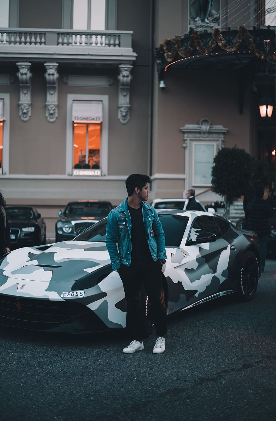 man in blue denim jacket and black pants stands beside parked green and white camouflage sports coupe, HD wallpaper