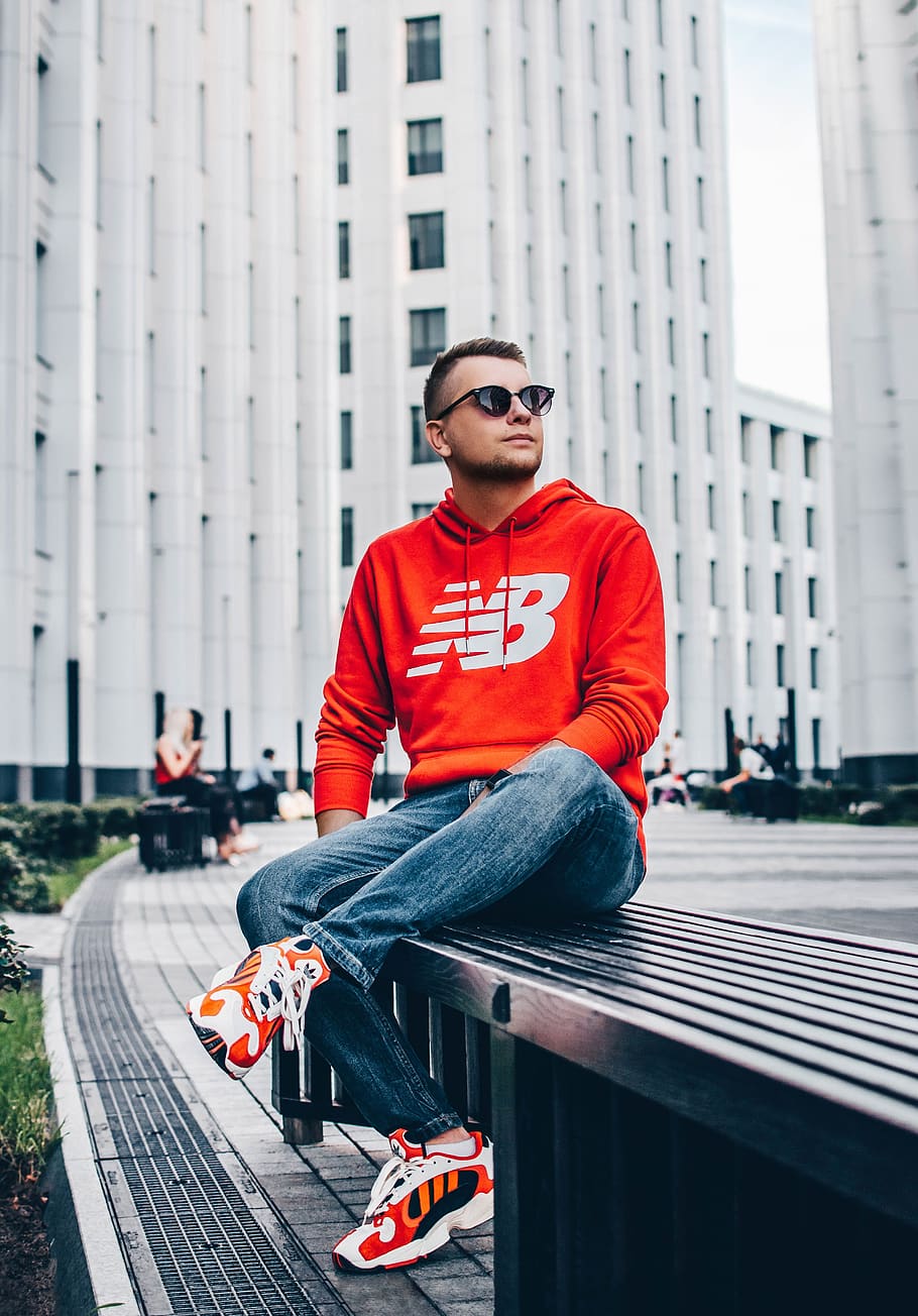man wearing red New Balance pullover hoodie sitting on black bench in front of white high-rise building, HD wallpaper