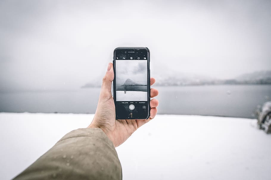 Photo of Person Wearing Brown Coat Holding Android Smartphone While Taking Picture of Mountain and Body of Water HD wallpaper
