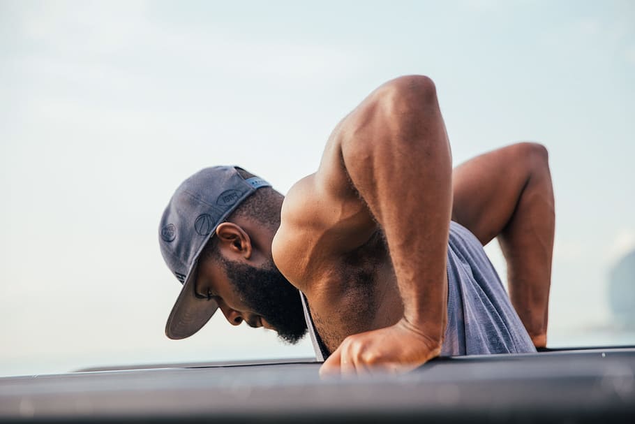 A bearded young African man wearing tank top and cap exercising on workout bars