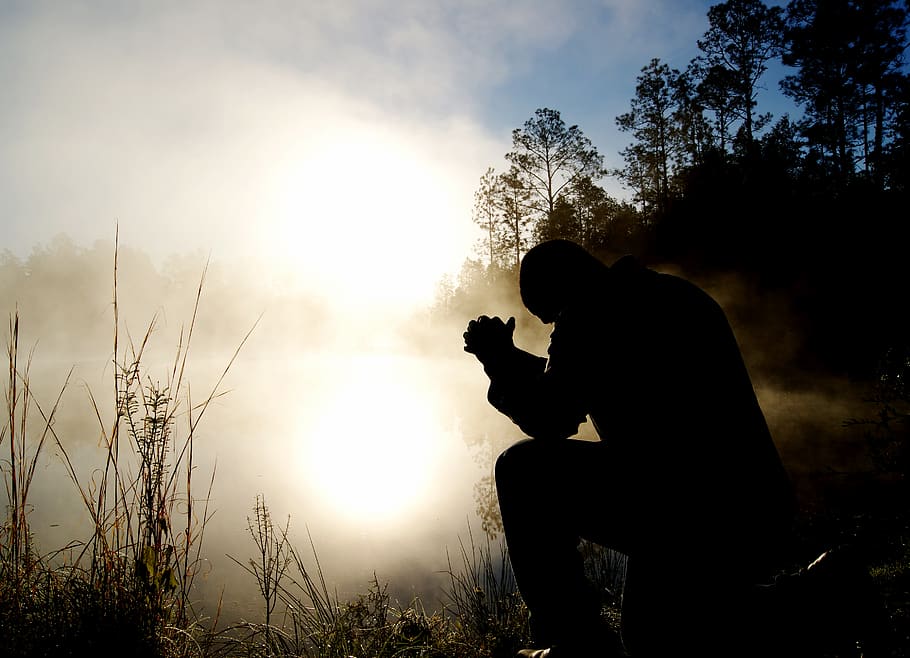 A silhouette of a man sitting outside with his hands together in prayer.