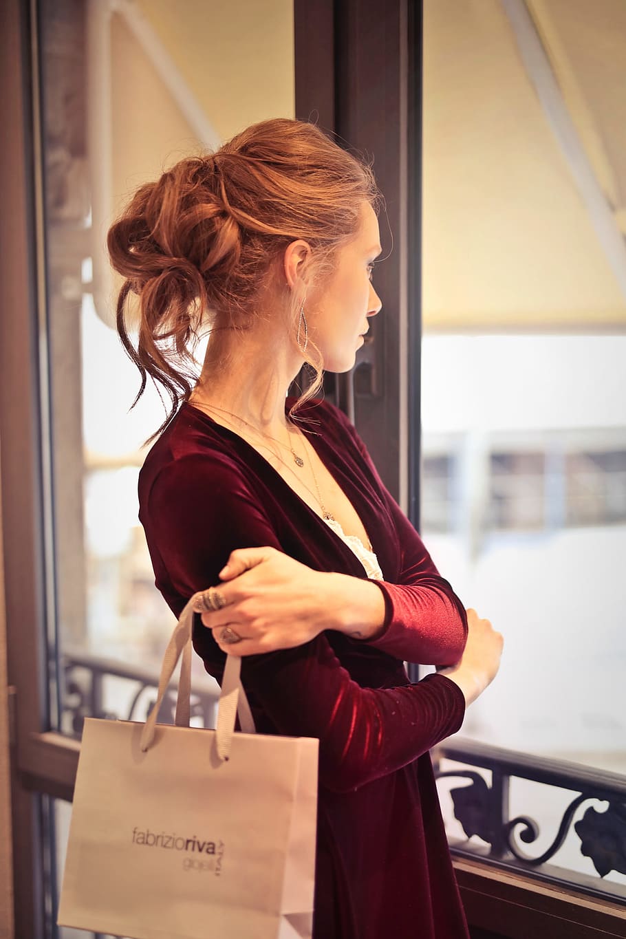 A young blond woman with elegant hair bun wearing a satin dress holding shopping bag in her hand looks out of the windows
