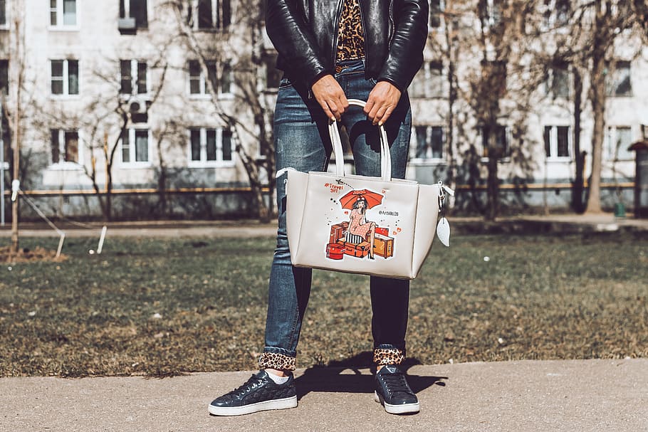 Person in Black Leather Jacket and Blue Denim Jeans Holding White Leather Tote Bag