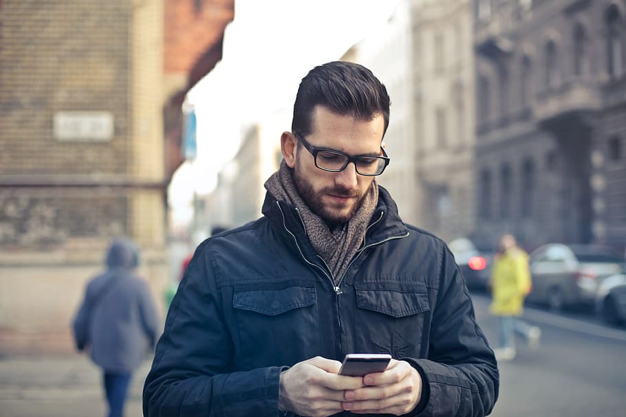 Young Man In Black Winter Jacket and Spectacles, Using His Smartphone While Walking On The Road