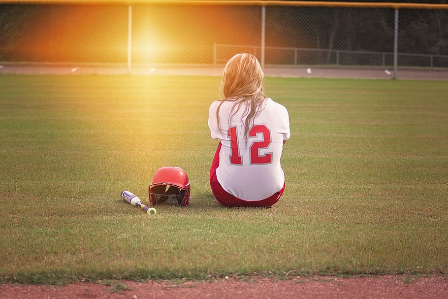 Female Baseball Player Sitting on Grass Field Beside Helmet and Baseball Bat