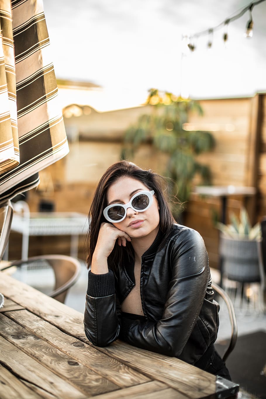 woman wearing black leather jacket and white framed sunglasses with right hand ion right cheek sitting beside brown wooden table, HD wallpaper
