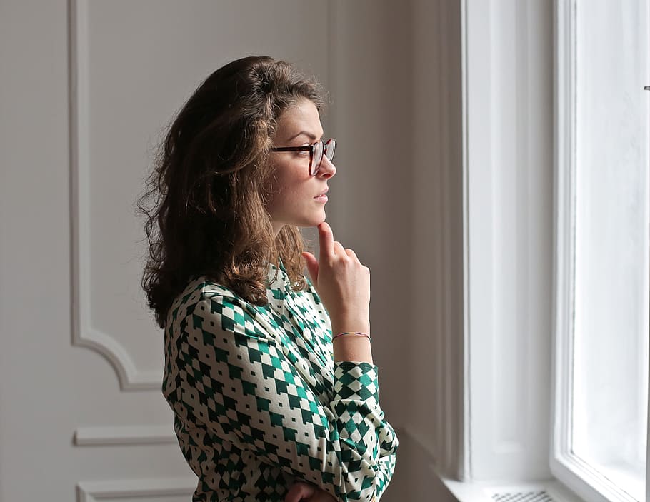 Young woman wearing printed collared shirt and spectacles keeping hand on chin and looking away while standing near window, HD wallpaper