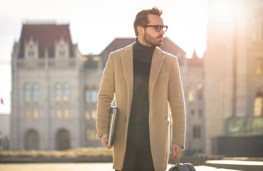 Young white bearded male wearing black turtleneck top with long overcoat and eyeglasses carrying a laptop,as he walks on the street