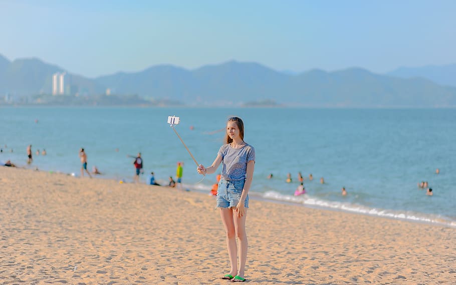 Woman Standing on Seashore Taking Selfie Using Monopod at Daytime HD wallpaper