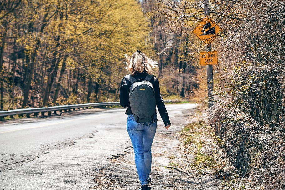 Woman in Blue Denim Fitted Jeans and Wearing Grey Backpack Walking on Gray Asphalt Road Near Road Signage and Trees at Daytime, HD wallpaper