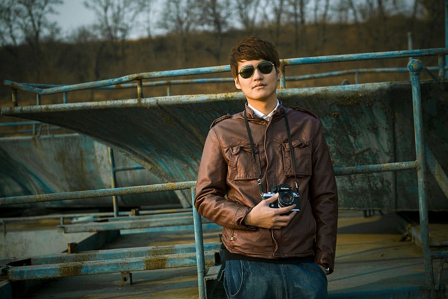 Man in Brown Leather Jacket Wearing Black Framed Sunglasses Holding Black Dslr Camera Standing in Front of Black Steel Bar during Daytime HD wallpaper