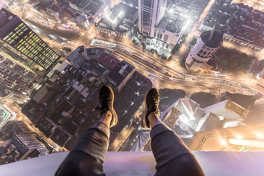 A person sitting on top of a skyscraper and dangling their legs over the ed...