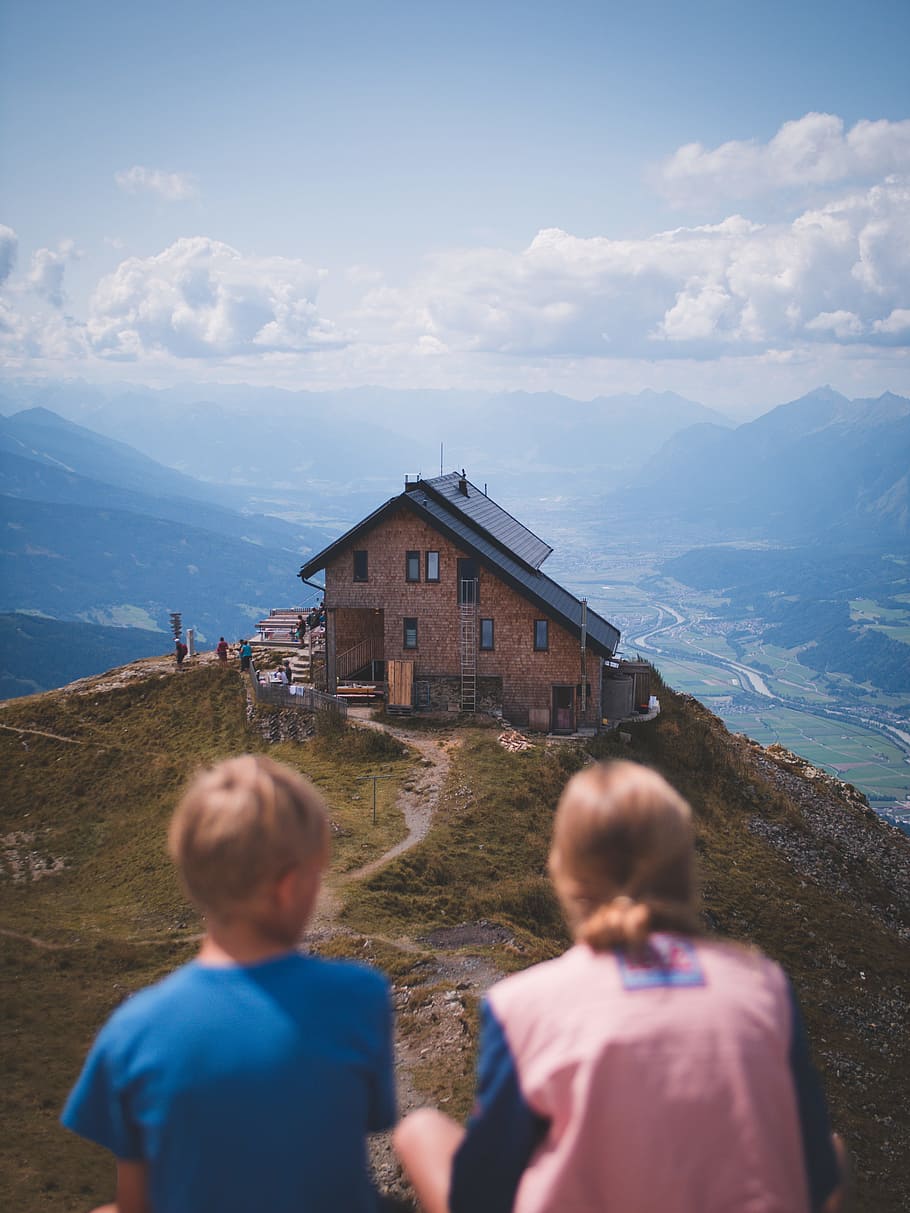 boy and girl sitting on cliff overlooking house at daytime, austria HD wallpaper