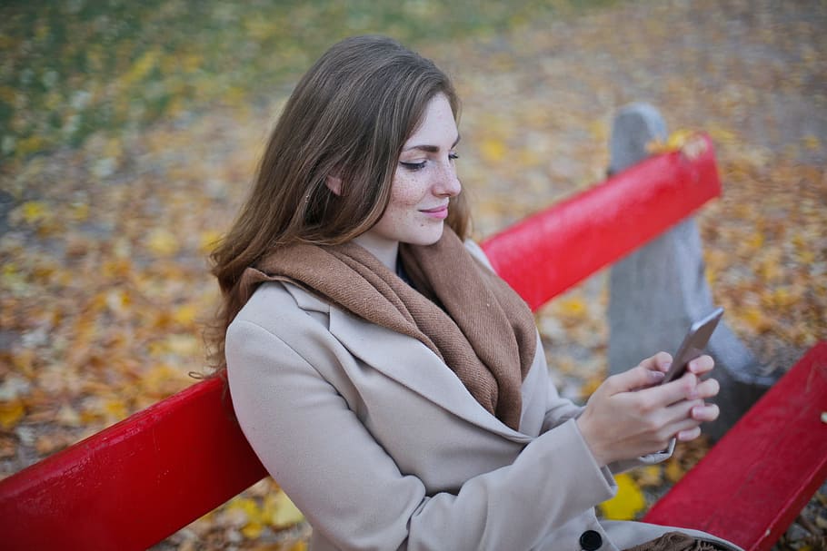 Young Woman Messaging On Her Mobile Phone While Sitting On Red Bench In Autumn Park HD wallpaper