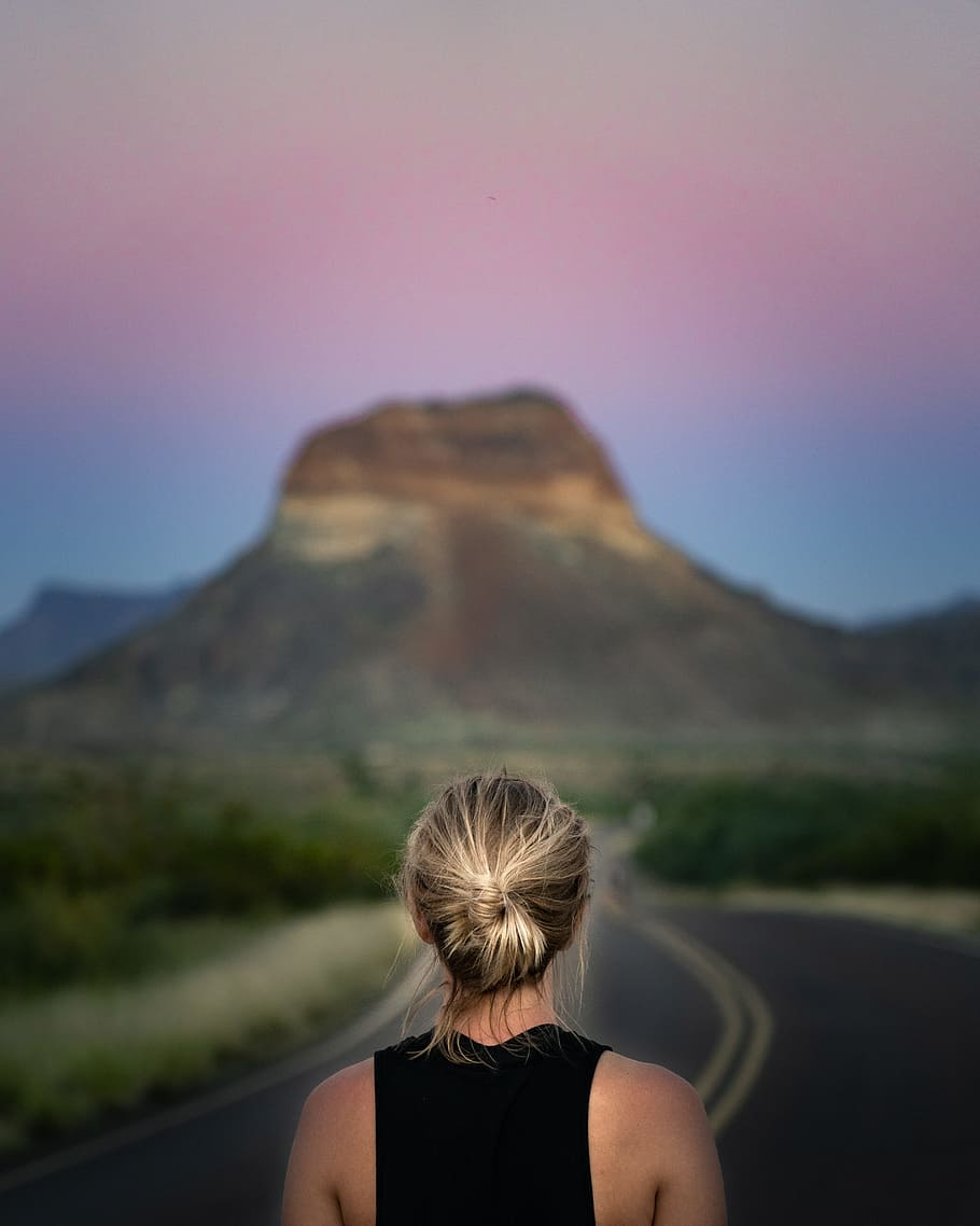 woman wearing black sleeveless top standing on road facing brown rock formation HD wallpaper