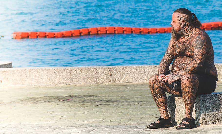 Man With Tattoos Sitting on Gray Concrete Floor Near Body of Water HD wallpaper