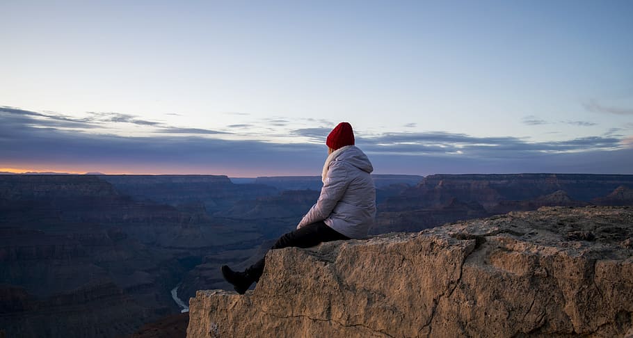 Person Wearing Gray Hooded Jacket and Black Pants Sitting on Mountain Cliff during Sunset HD wallpaper