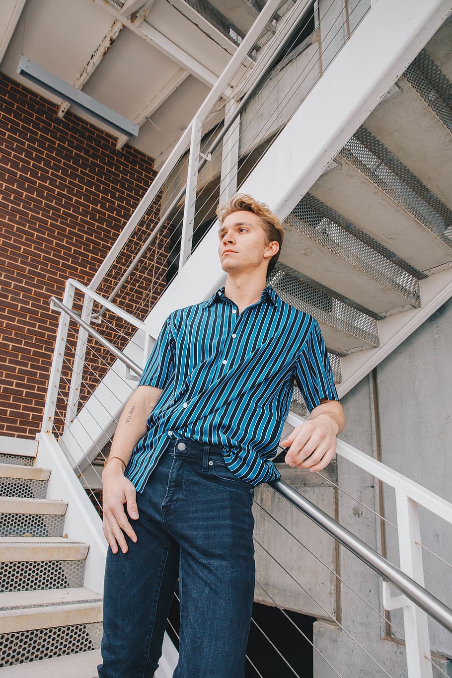 man in blue and black striped button up polo shirt stands on stairway leaning on balustrade
