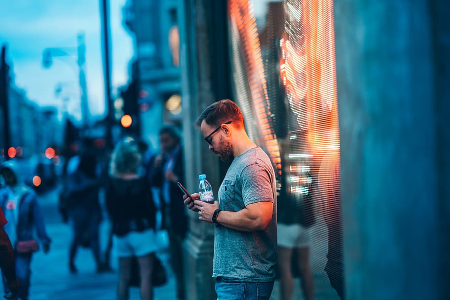 man standing beside wall holding smartphone and water bottle, HD wallpaper