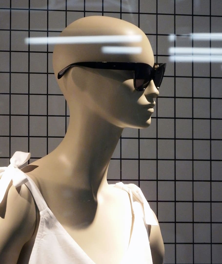 The bald head of a female mannequin wearing a pair of dark sunglasses against a background of strobe lighting reflecting in a shop window