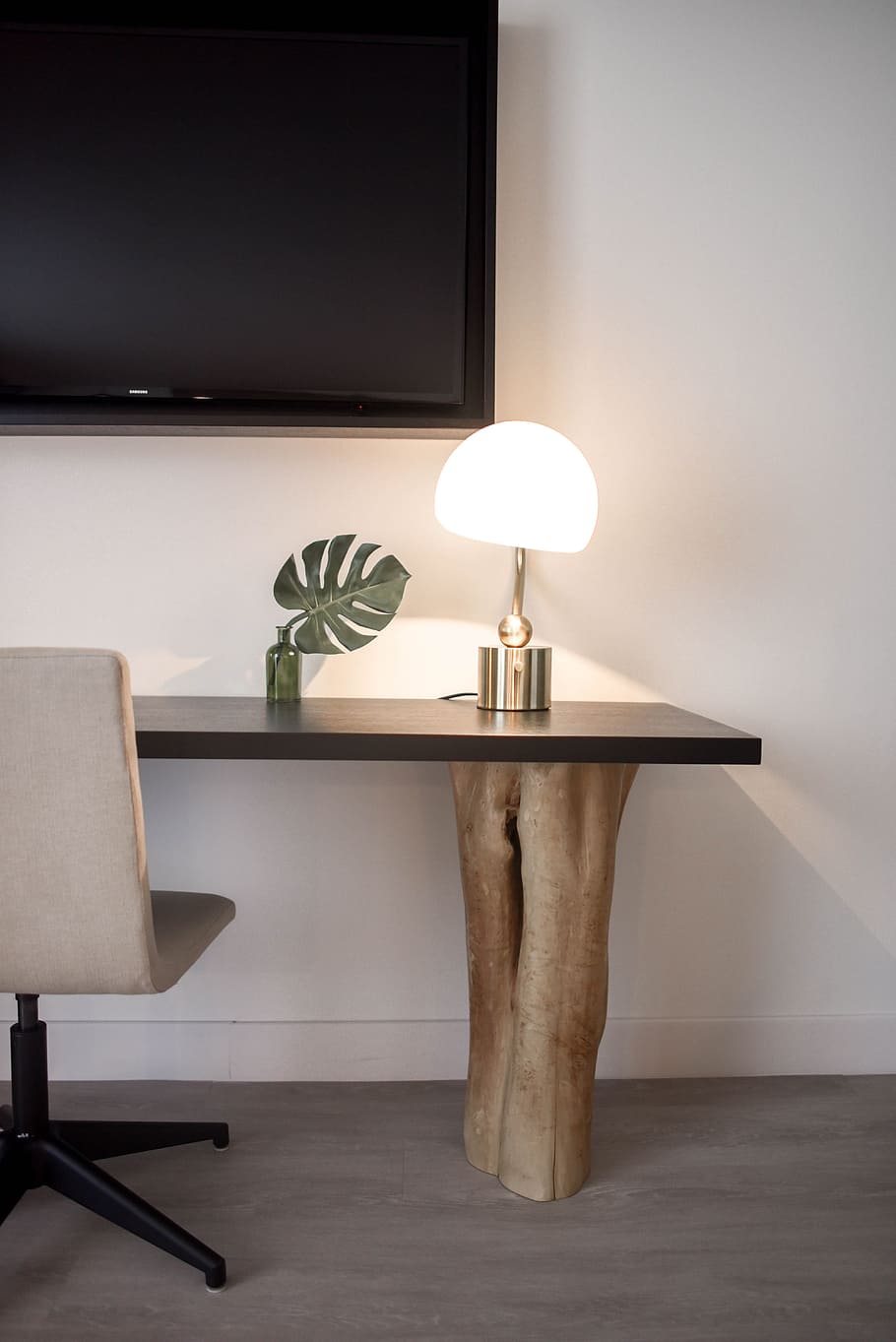 Stainless Steel Base White Shade Table Lamp on Brown Wooden Desk Near White Painted Wall With Wall Mounted Flat Screen T V