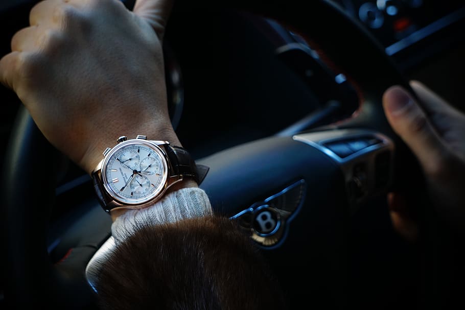 person wearing round gold-colored framed chronograph watch holding black vehicle steering wheel inside car