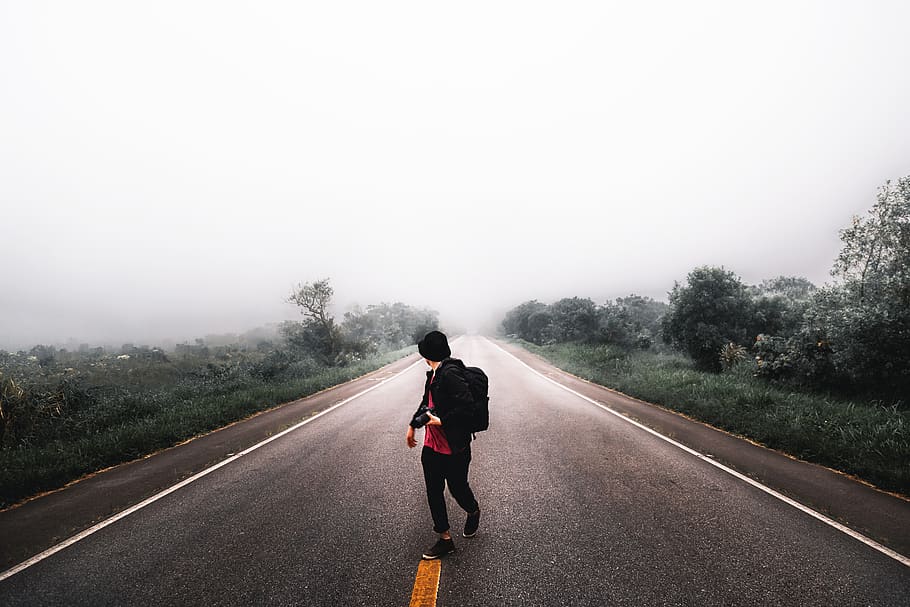 Person Waling in the Middle of Road in Between Grass Field Under White Skies HD wallpaper