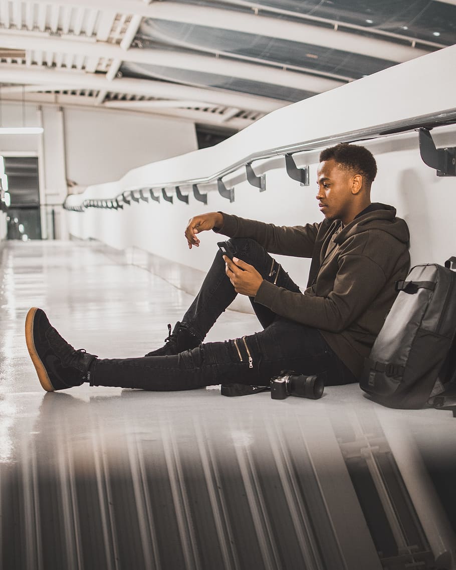 man wearing black jacket sitting while leaning on wall and using smartphone HD wallpaper