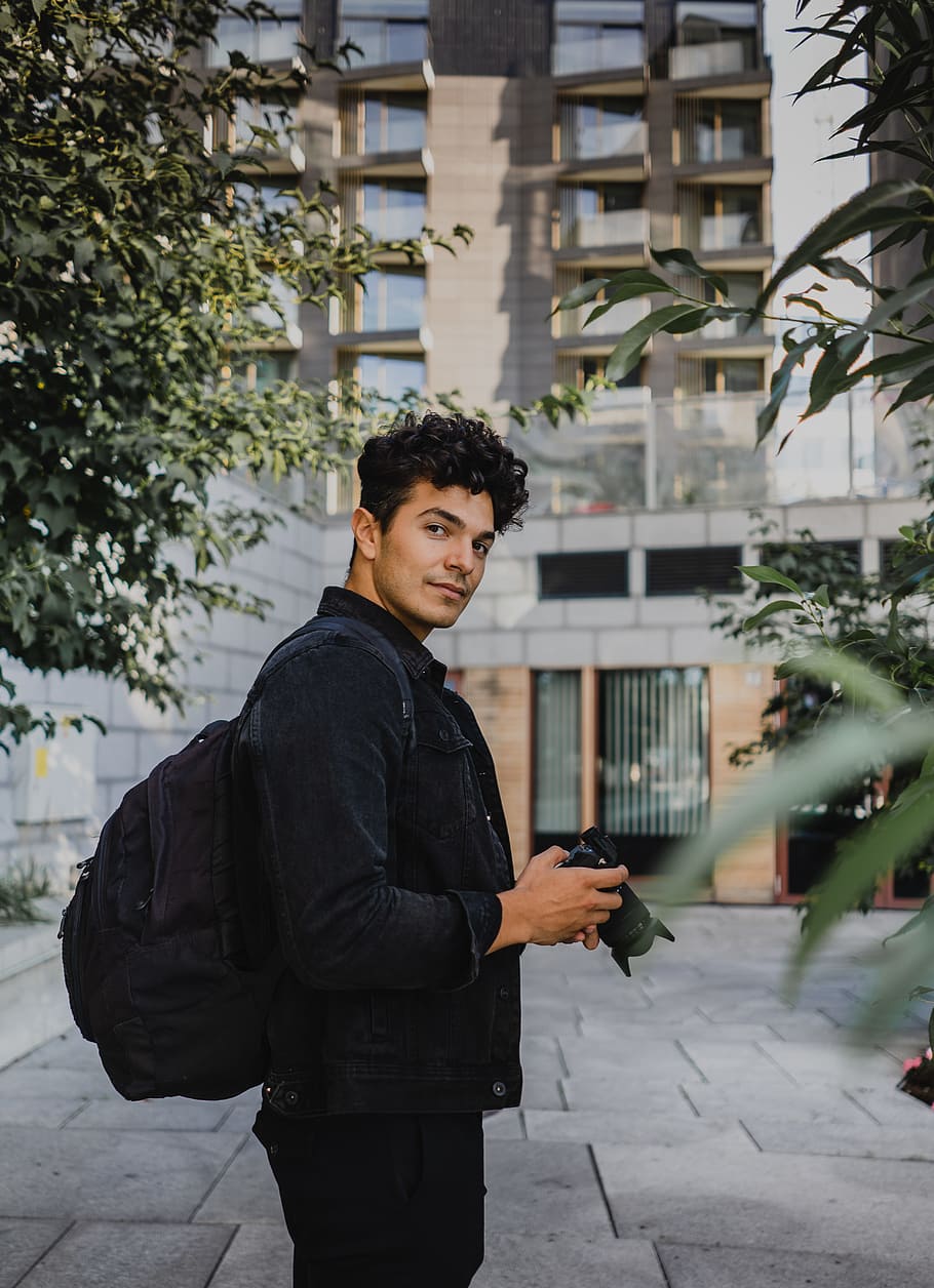 man holding DSLR camera carrying backpack near building, male HD wallpaper