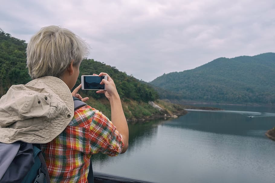 Person Holding Smartphone While Taking Photo of Calm Water Near Mountain at Daytime HD wallpaper