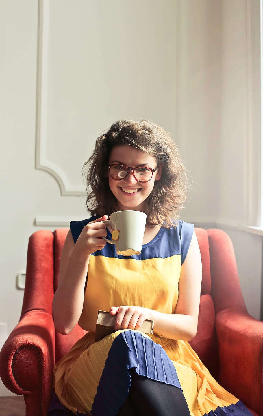 Young Woman Wearing Yellow And Blue Dress With Spectacles, And Holding A Book While Enjoying A Cup Of Tea At Home