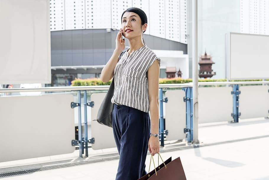 Woman Wearing Brown and White Pinstriped Sleeveless Top Holding Paper Tote Bags Using Smartphone Near White Buildings
