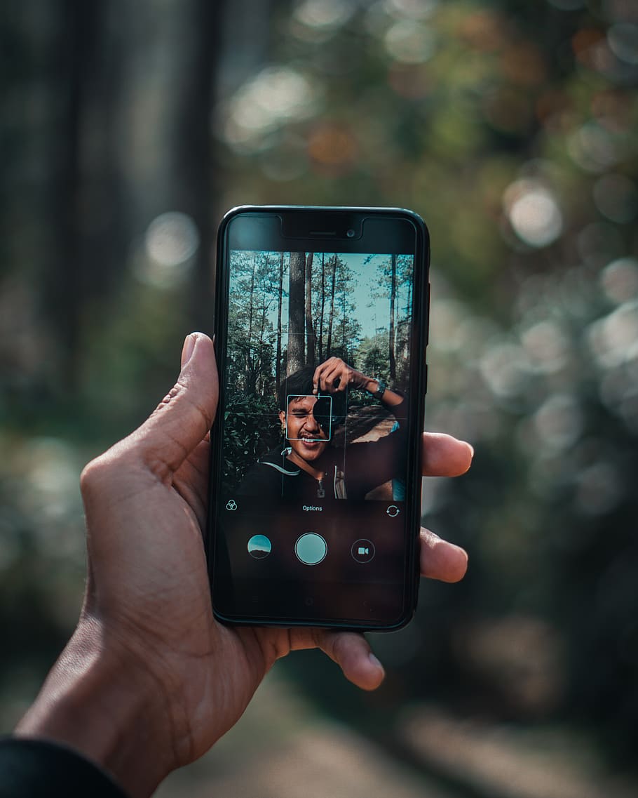 person holding black smartphone while taking selfie under the tree
