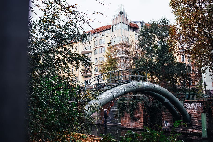 A curved metal pipeline with walkway railing over an urban river surrounded by buildings and trees HD wallpaper