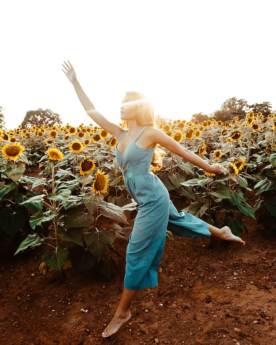 woman wearing green sleeveless rompers standing and waving hands near garden of sunflower at daytime